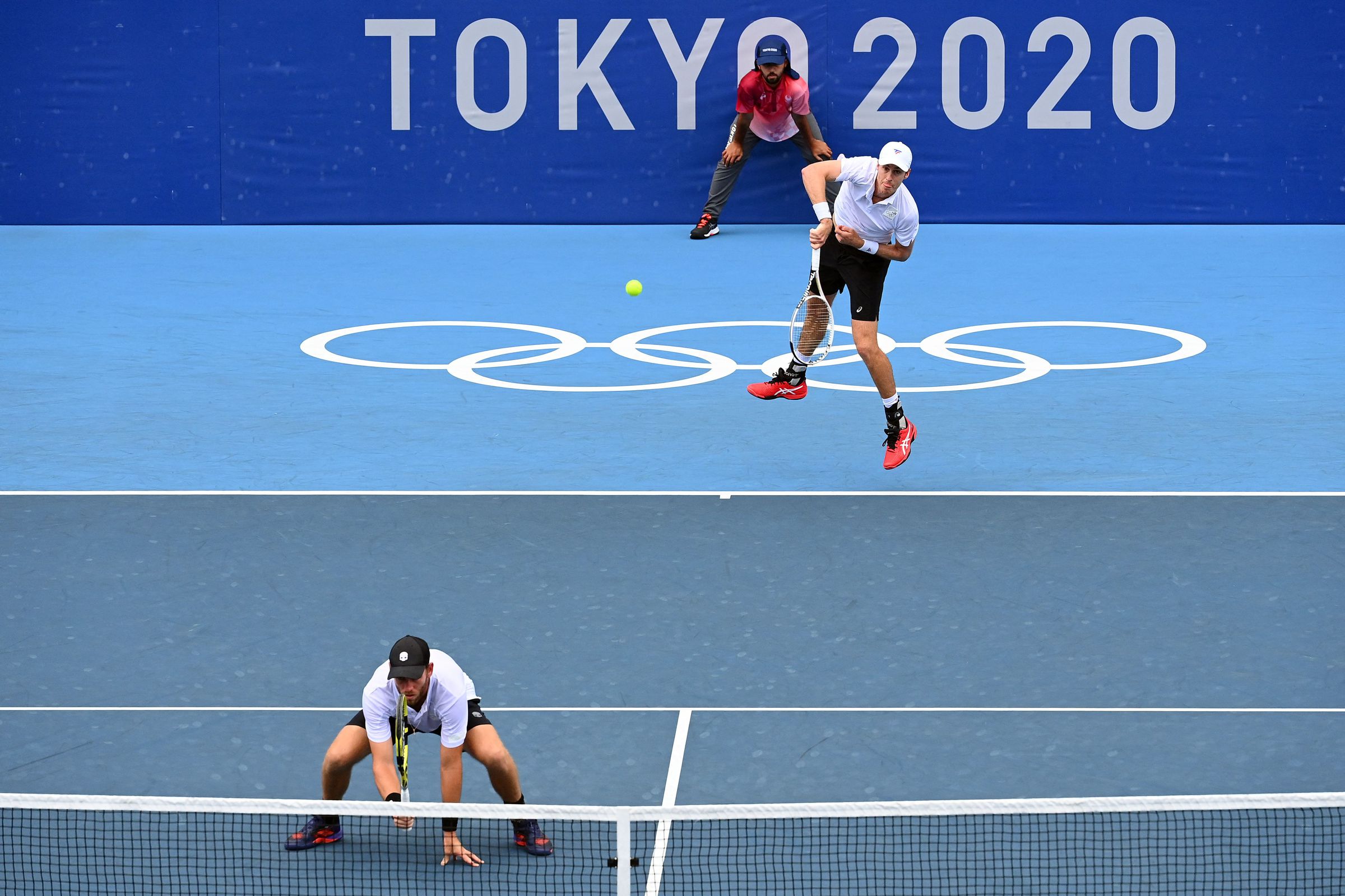 A man leaps to hit a tennis ball on an Olympic court. Another man is crouching on the court in front of him.