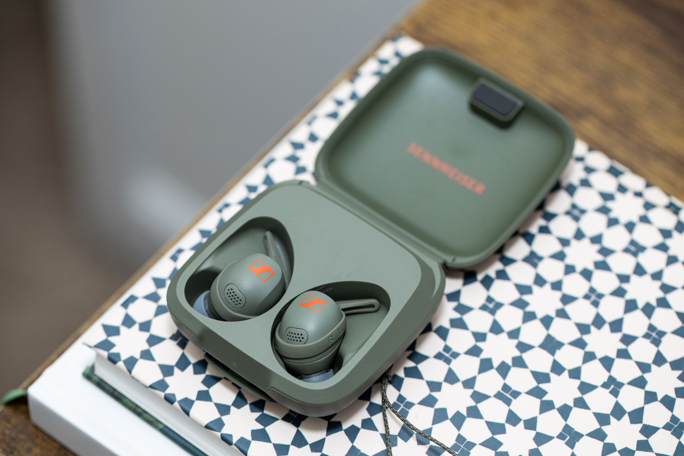 Olive-colored Sennheiser Momentum Sport buds in their case on top of a patterned book.