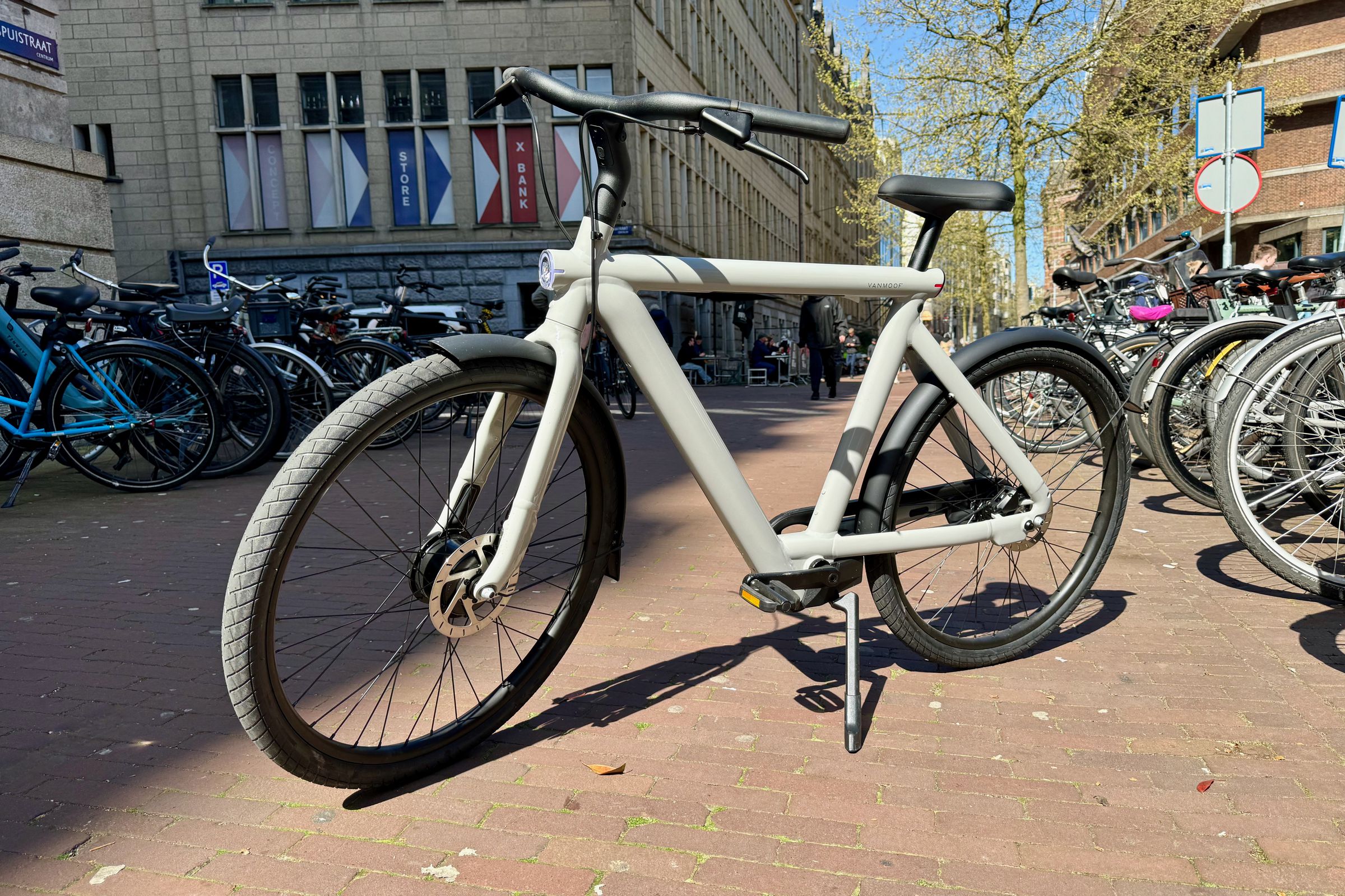 A light grey VanMoof S5 e-bikes sits on a brick sidewalk in Amsterdam between two long rows of bicycles.