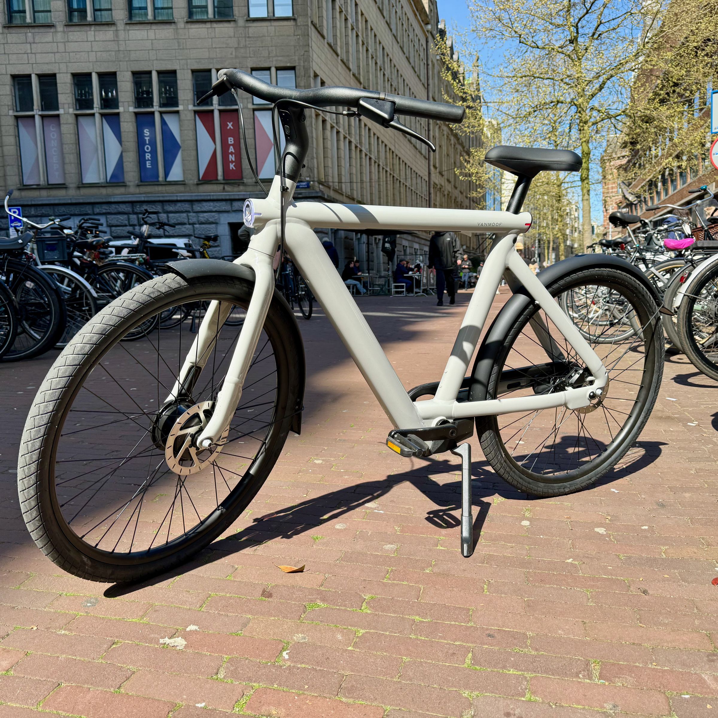 A light grey VanMoof S5 e-bikes sits on a brick sidewalk in Amsterdam between two long rows of bicycles.