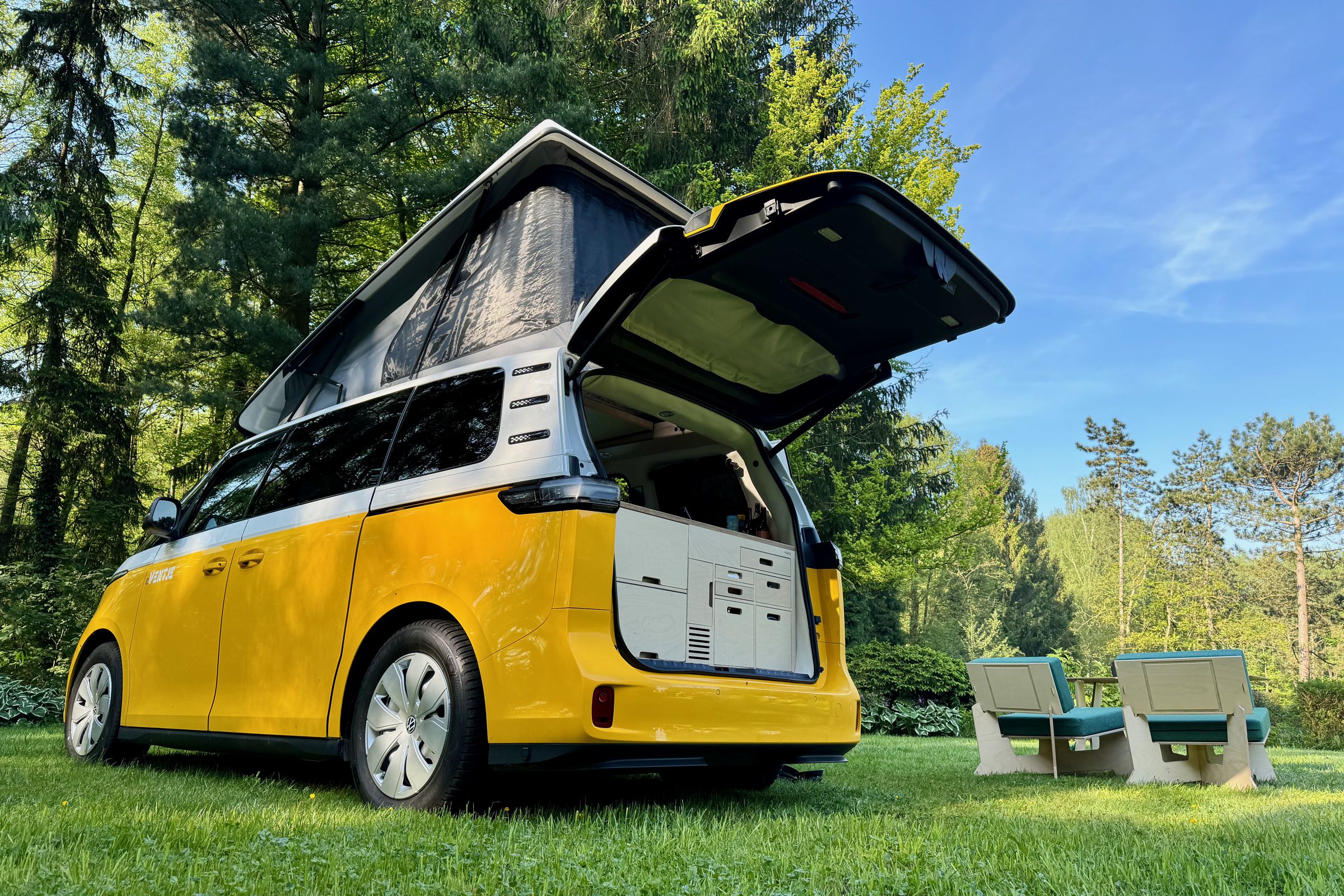 A customized ID Buzz Ventje sits on the grass beneath the trees with the pop-top and tail gate open and furniture setup on the lawn.