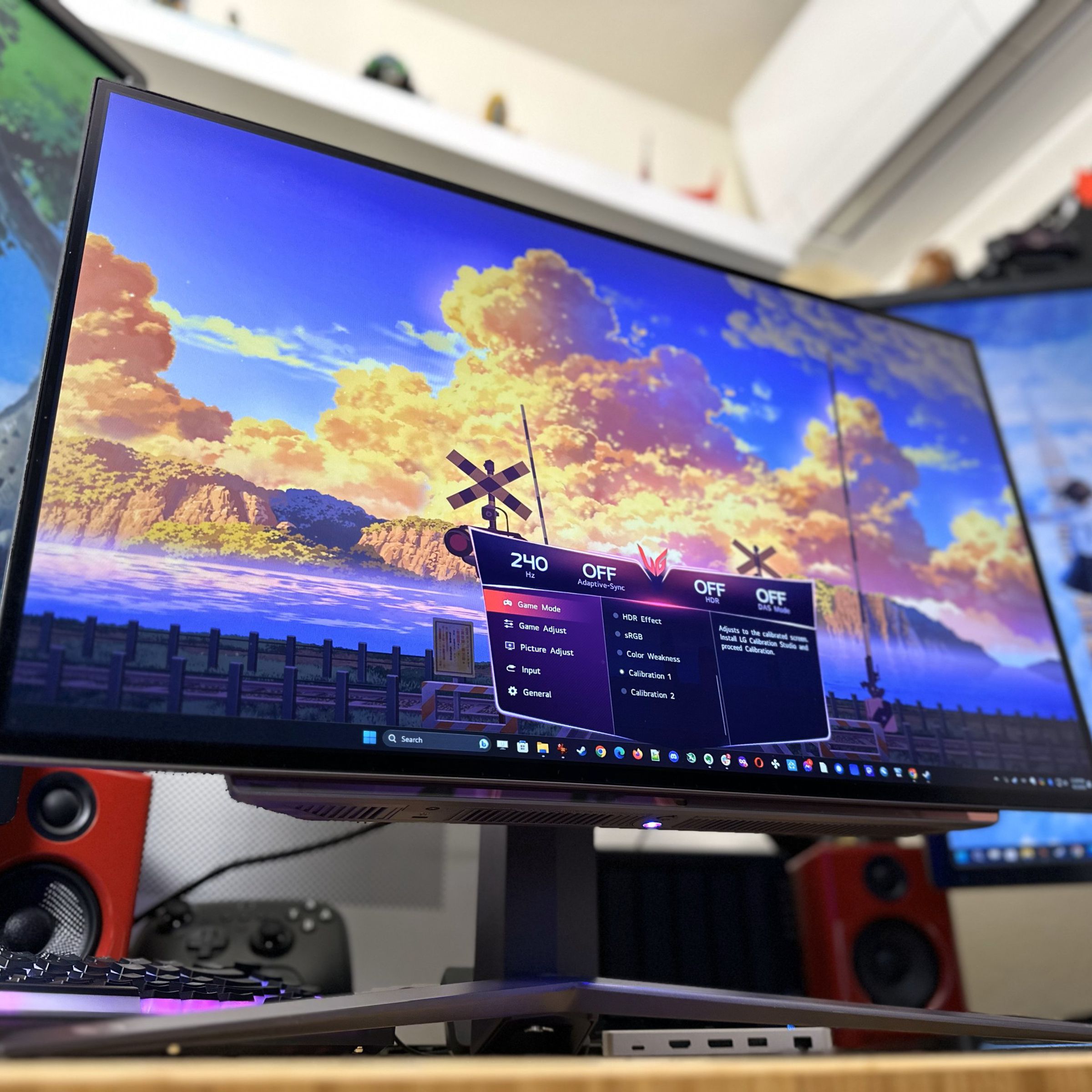 A bright, colorful OLED monitor with a big V-shaped stand underneath, flanked by two other monitors in portrait mode. The main monitor has a colorful sky on screen with big orange sunset-lit clouds and an on-screen display showing it’s running at 240Hz.